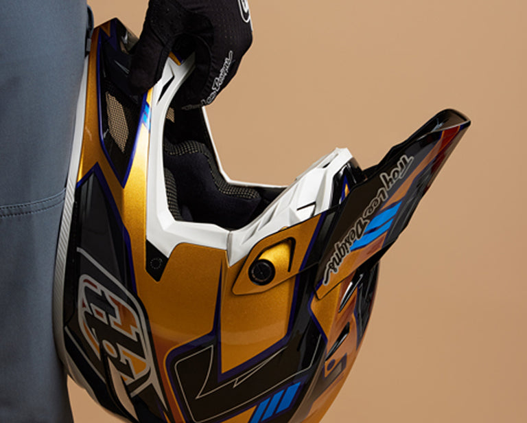 A rider holding a Troy Lee Designs full face helmet in front of a brown background